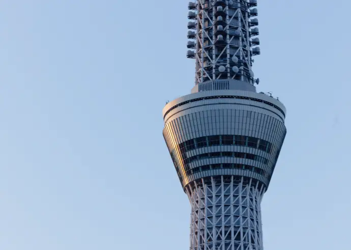 晴天の下、屋上で遠くの東京スカイツリーを背景に作業を行う、青い作業服の人物。
