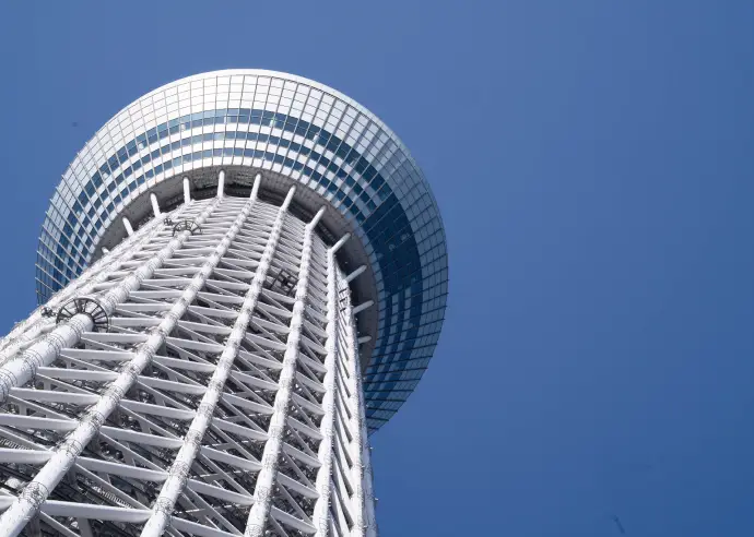 青空を背景に、天望デッキ付近の鉄骨フレームと丸みを帯びた塔体上部を斜め下から見上げた写真。