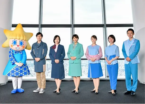 東京スカイツリーの展望台を背景に並ぶ、公式キャラクターのソラカラちゃんと、デザインの異なる複数種類のユニフォームを着用した6名のスタッフ。
