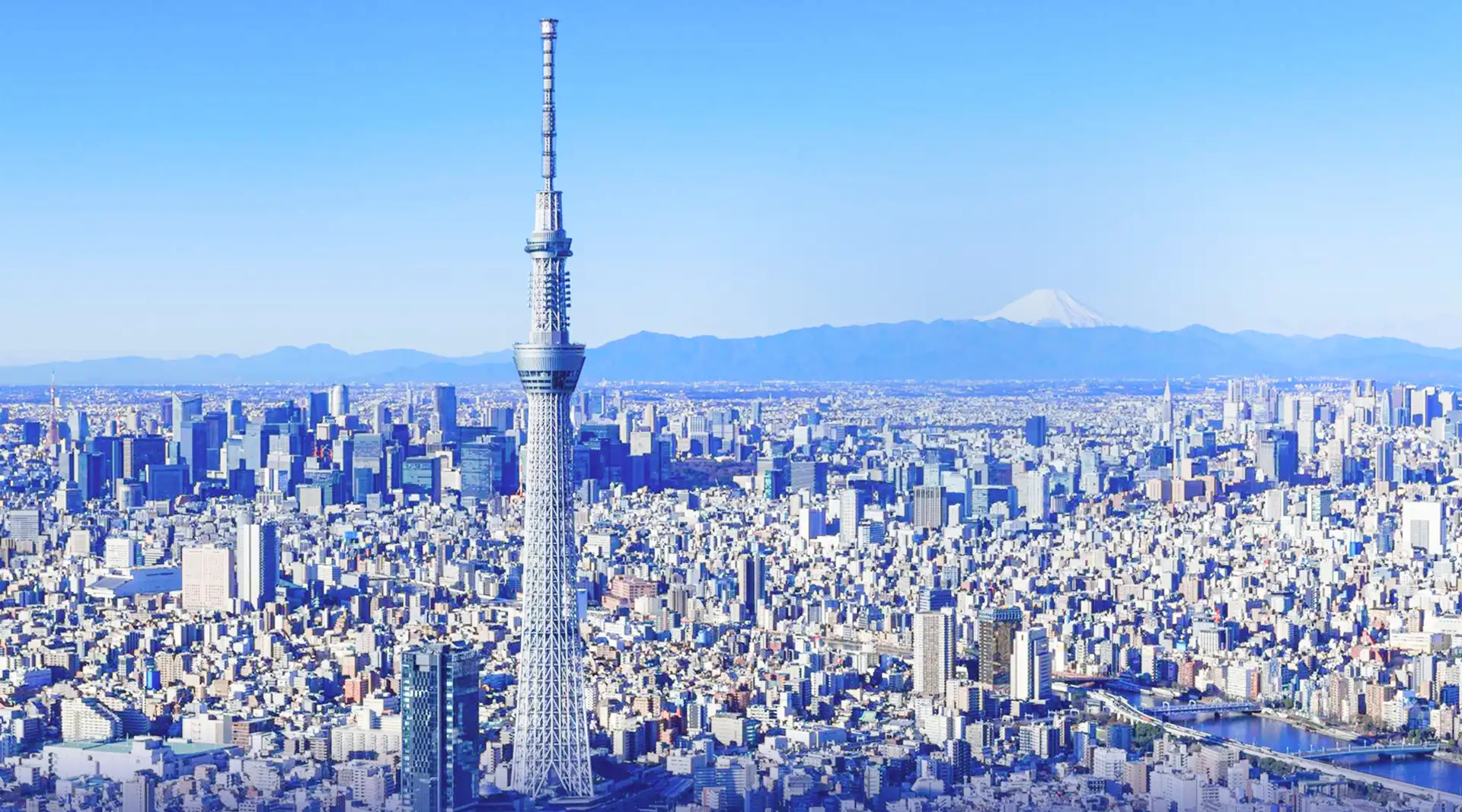 東京の街並みと東京スカイツリーを遠景で捉え、奥に富士山が見える空撮風景