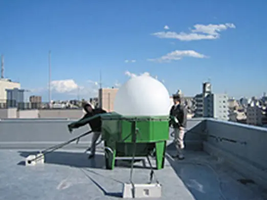 屋上で白い球体状の気球を機材に取り付け、放球準備をする作業員の様子