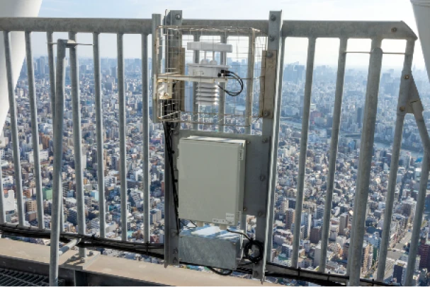 都市のパノラマを背景に、高層ビルの手すりに取り付けられた気象観測装置と通信用機器