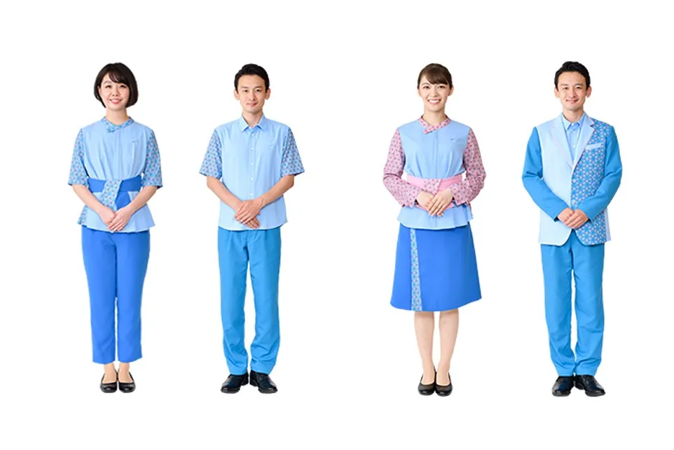 Uniform worn by Ticket Counter and guidance Staff . From left to right, there are four people lined up: a woman in pants, a man in a short-sleeved shirt, a woman in a skirt, and a man in a jacket. All of them feature a modern Japanese design with a Sky blue base and cherry blossom pink Japanese patterns on the sleeves, sashes, and collars.