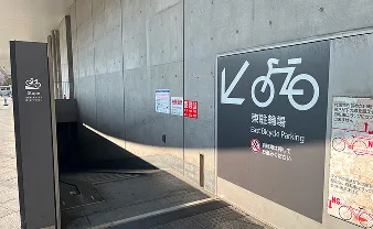 東駐輪場の入り口。コンクリートの壁に自転車のピクトグラムと「東駐輪場 East Bicycle Parking」の文字があり、地下へ続くスロープと階段が設置されている。