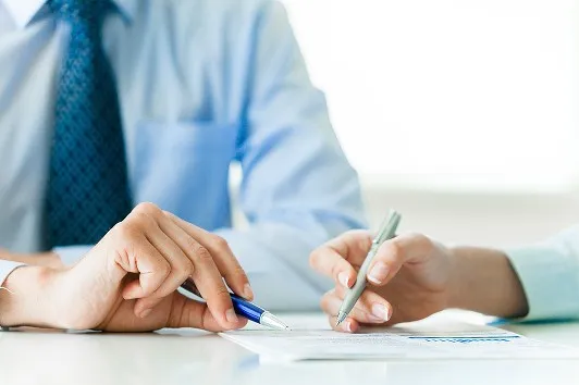 ビジネスウェアを着た二人が、書類を前にペンを手にしている手元のアップ。