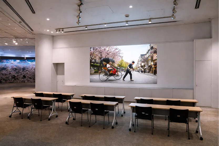 壁のスクリーンに人力車の写真が投影されている、セミナー形式に設営された会場。手前には黒い椅子と長机が並んでいる。