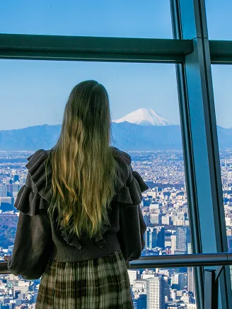 展望台の窓越しに、遠くの富士山を眺める後ろ姿の女性。