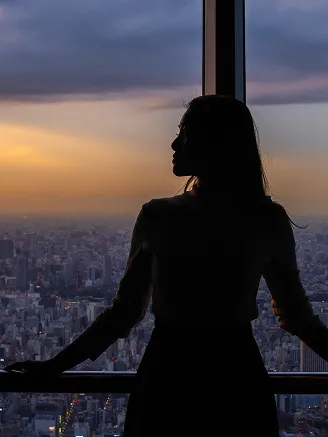 夕景の窓際に立つ女性のシルエット。逆光の中で都会の夕暮れを見つめる様子。