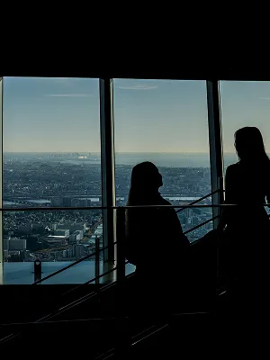展望台の窓際に座り、夜景を眺めながら語り合う二人のシルエット。室内は暗く、窓の外には宝石を散りばめたような街の光が見える。
