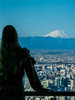 展望台から、夕日に照らされた富士山のシルエットを眺める女性の背中。眼下には影になった街並みが広がっている。