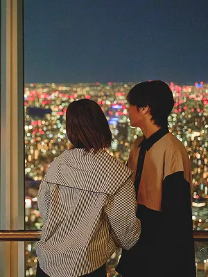 夜の展望台で楽しそうに会話する男性と女性。背景には、ぼんやりと光り輝く街の夜景が広がっている。
