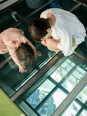 ガラス張りの床の上に寝そべり、真下の景色を覗き込む子どもと大人。高層階からのスリリングな視界を楽しんでいる様子。