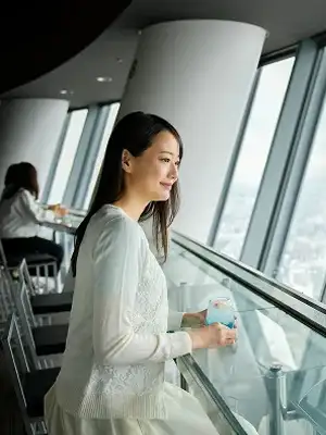 展望台の窓際に立ち、外を眺める白い服を着た女性。柔らかい光が差し込み、穏やかな表情で景色を楽しんでいる。