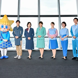 東京スカイツリーの展望台を背景に並ぶ、デザインの異なる複数種類のユニフォームを着用したスタッフ。