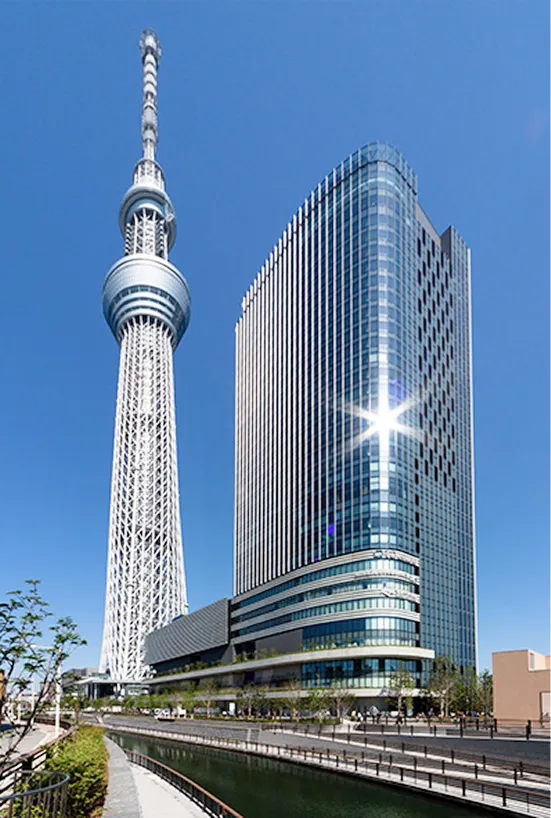 青空の下、川沿いにそびえ立つ東京スカイツリーと、その隣に隣接する高層ビル「東京スカイツリーイーストタワー」の全景。手前には北十間川と遊歩道が見える。