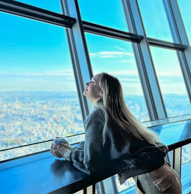 展望台の窓から青空と街並みを眺めるドリンクを持った女性