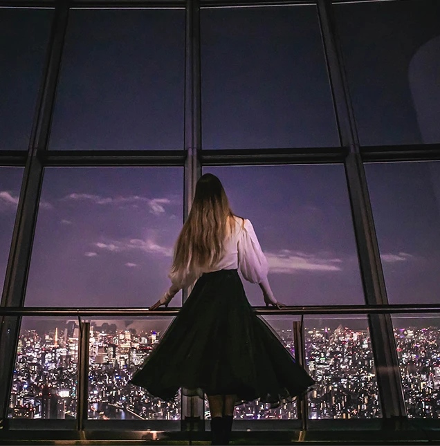 夜景を背景に展望台の窓際に立つ黒いスカート姿の女性の後ろ姿