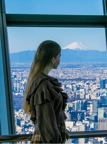 展望台の窓から雪化粧した富士山と東京のビル群を眺める女性の横顔