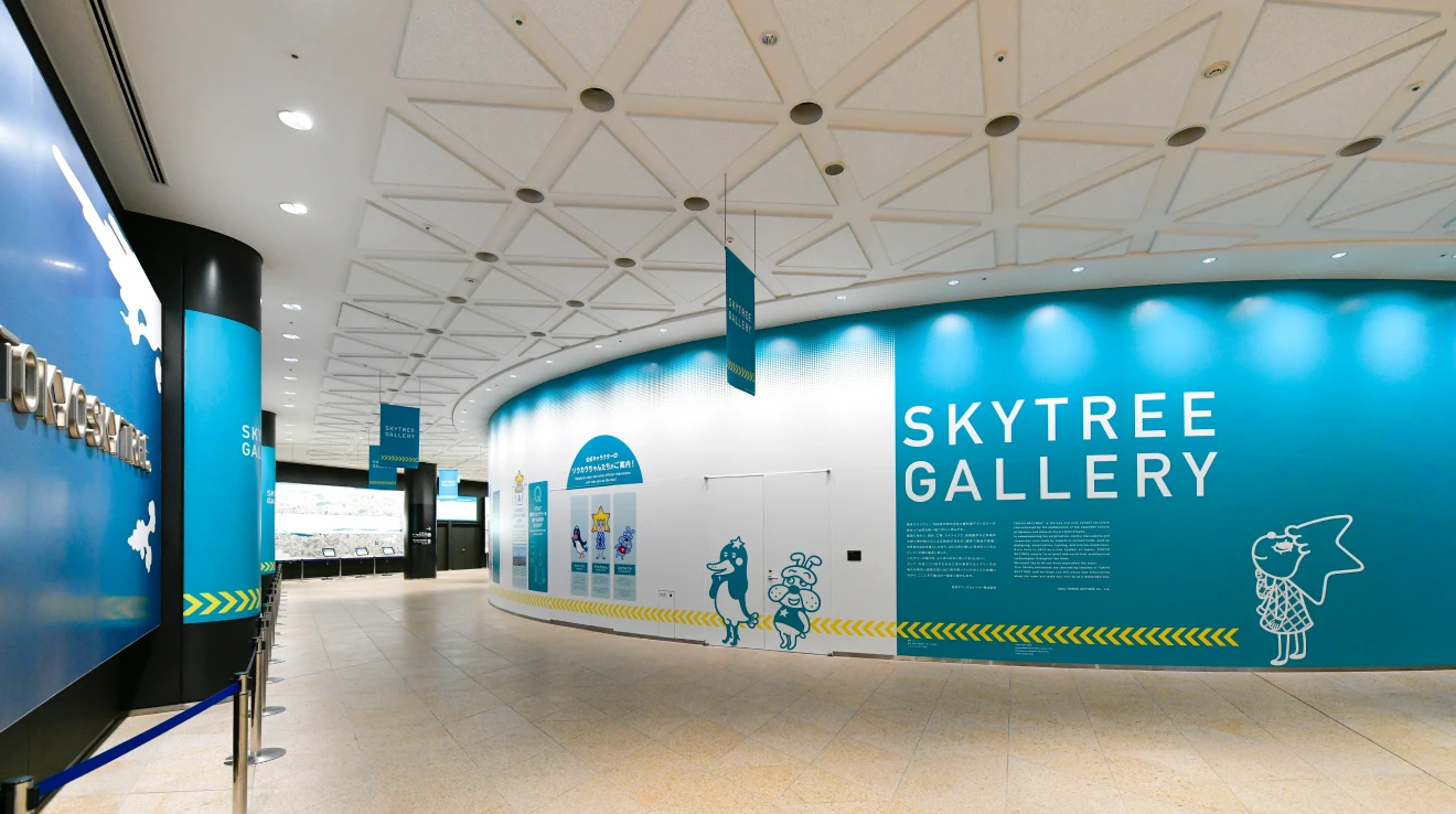 A view of the entrance to the TOKYO SKYTREE Gallery. The exhibition space features a vibrant light blue wall adorned with the Official characters"Sorakara-chan" and the logo.