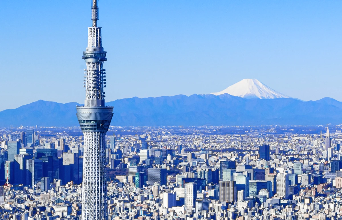 東京スカイツリーの背後に、雪を冠した富士山が遠くそびえ立つ冬の風景。手前にはどこまでも続く東京の広大な街並みが広がっている。