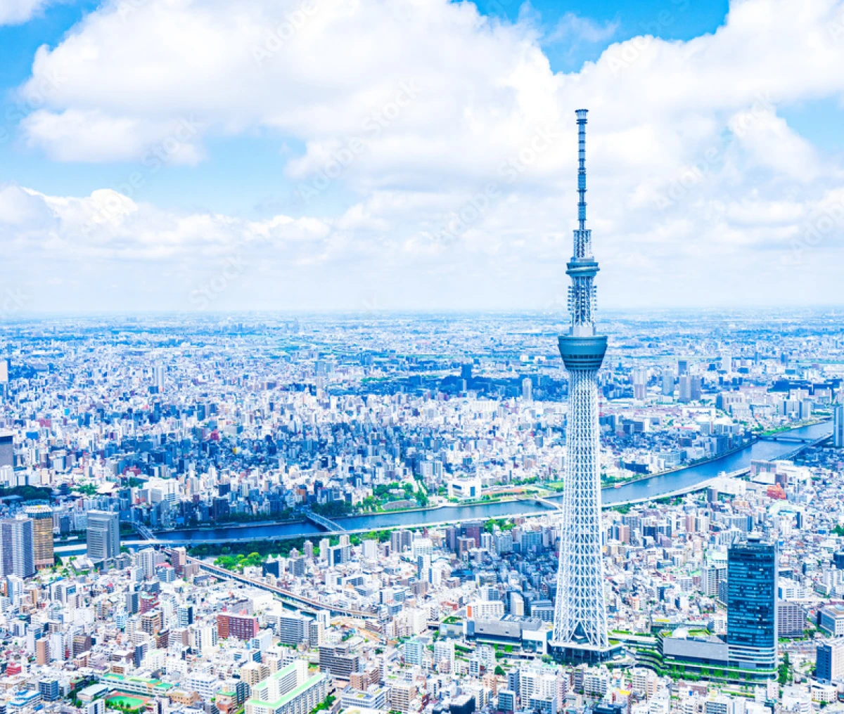 白い雲が浮かぶ青空の下、東京スカイツリーを中心に捉えた都市の俯瞰写真。隅田川が街の間を流れ、光り輝く街並みが地平線まで続く開放的なカット。