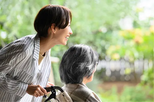 緑の見える屋外で、車椅子に乗った高齢の女性を笑顔でサポートする若い女性