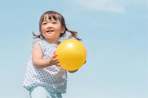 青空の下で、白いドット柄の服を着た幼い女の子が黄色いボールを持って笑顔で走っている様子
