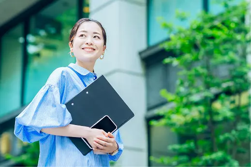 青いストライプのシャツを着てバインダーとスマートフォンを抱え、屋外で微笑む女性社員