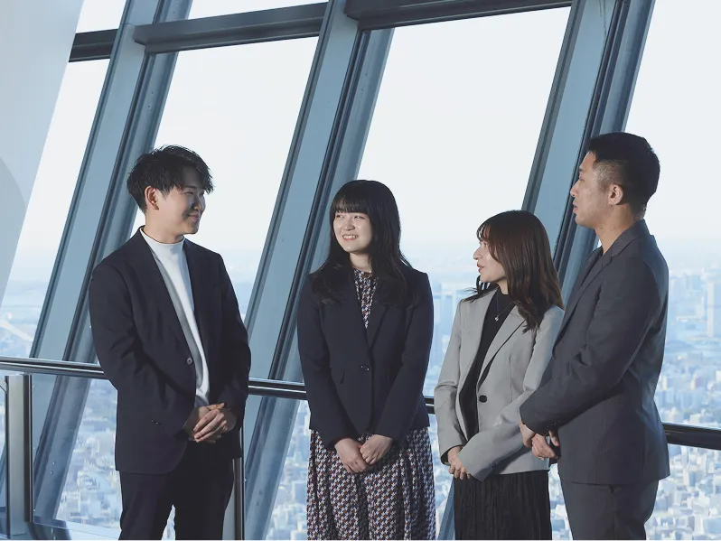 明るいオフィスの窓際で、笑顔で談笑する4人のビジネスパーソン