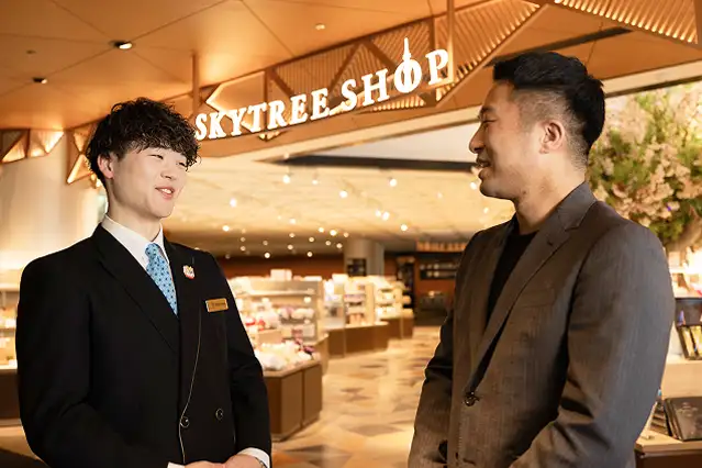 東京スカイツリーショップの入り口で、スーツ姿の男性スタッフが笑顔で会話している様子。