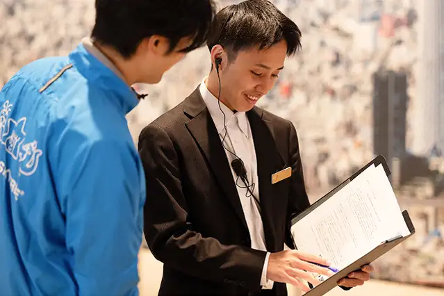 スーツ姿の男性スタッフがクリップボードを手に持ち、青いウェアを着たスタッフと内容を確認し合っている様子。