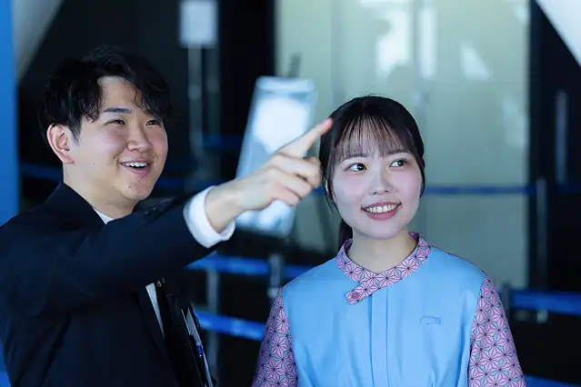 スーツ姿の男性スタッフが、青い制服を着た女性スタッフの隣で前方を指差し、案内や説明をしている様子。
