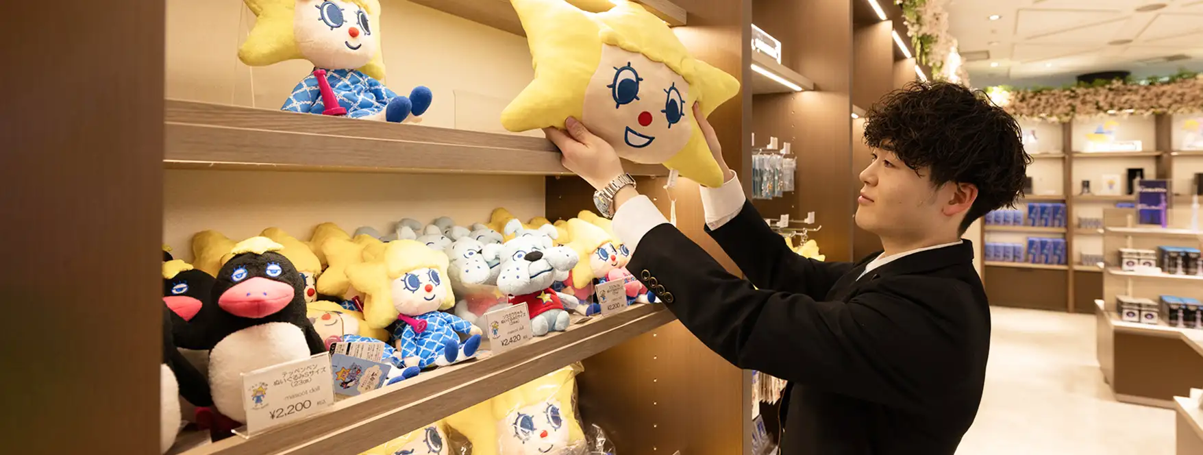 東京スカイツリー公式ショップにて、スーツ姿の男性スタッフが棚にあるソラカラちゃんのぬいぐるみを整えている様子。