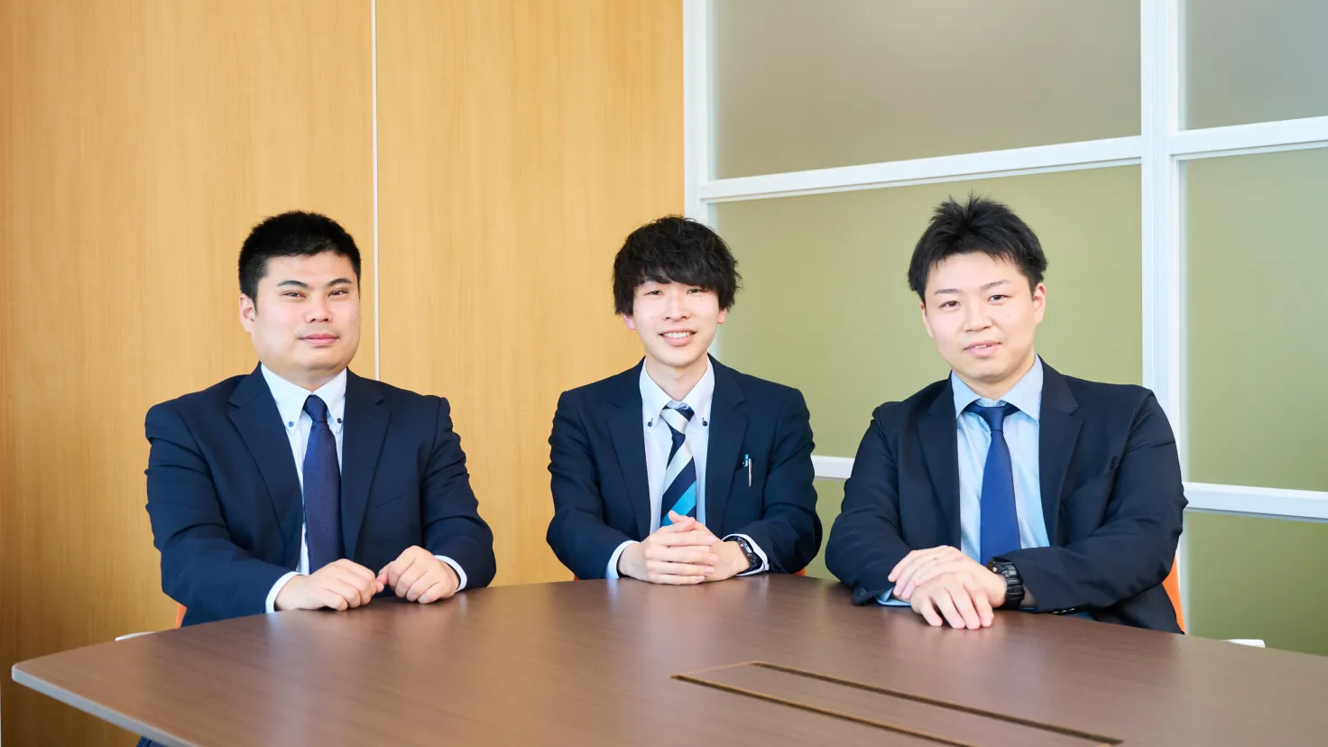 会議室のテーブルに座りカメラに向かって微笑むスーツ姿の社員3名