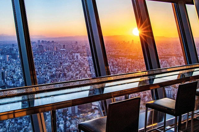 夕日に染まる東京の街並みを地上340メートルから一望できる、展望デッキのカフェカウンター。大きな窓に面した透明なカウンターテーブルに黒いハイチェアが並び、目の前には沈みゆく太陽と果てしなく広がる都会のパノラマが広がっています。