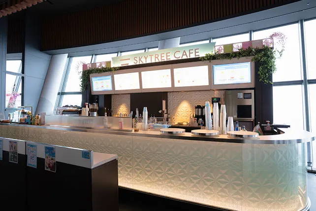 鮮やかなブルーのライトアップが施された、スタイリッシュな「SKYTREE CAFE」のカウンター。上部のモニターにはカラフルなドリンクや軽食のメニューが映し出されています。