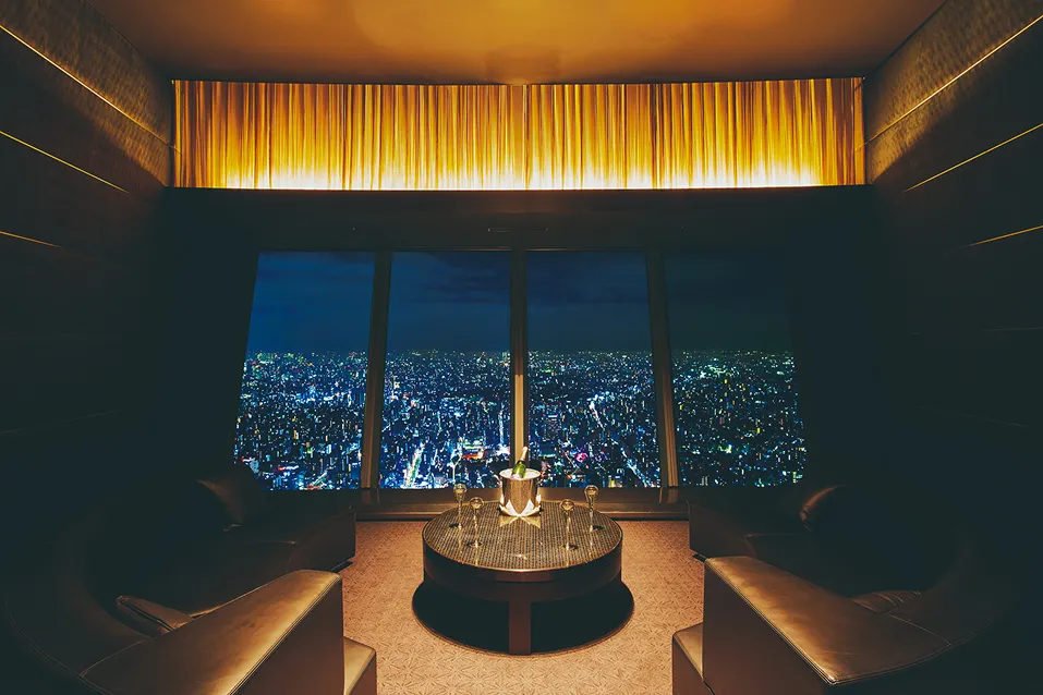 東京スカイツリーの展望デッキにあるレストランの個室。正面の大きな窓一面に、地平線まで続くまばゆい街の夜景が広がっています。室内は落ち着いた暖色の照明に包まれ、中央の円卓にはシャンパンボトルとグラスが用意され、特別なひとときを演出しています。