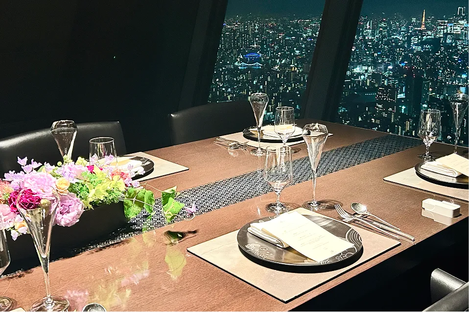 夜景を一望するレストランの個室。シックな木目調のテーブルには、色鮮やかな花々が飾られ、上品な和文様のプレートやシャンパングラスが美しくセッティングされています。