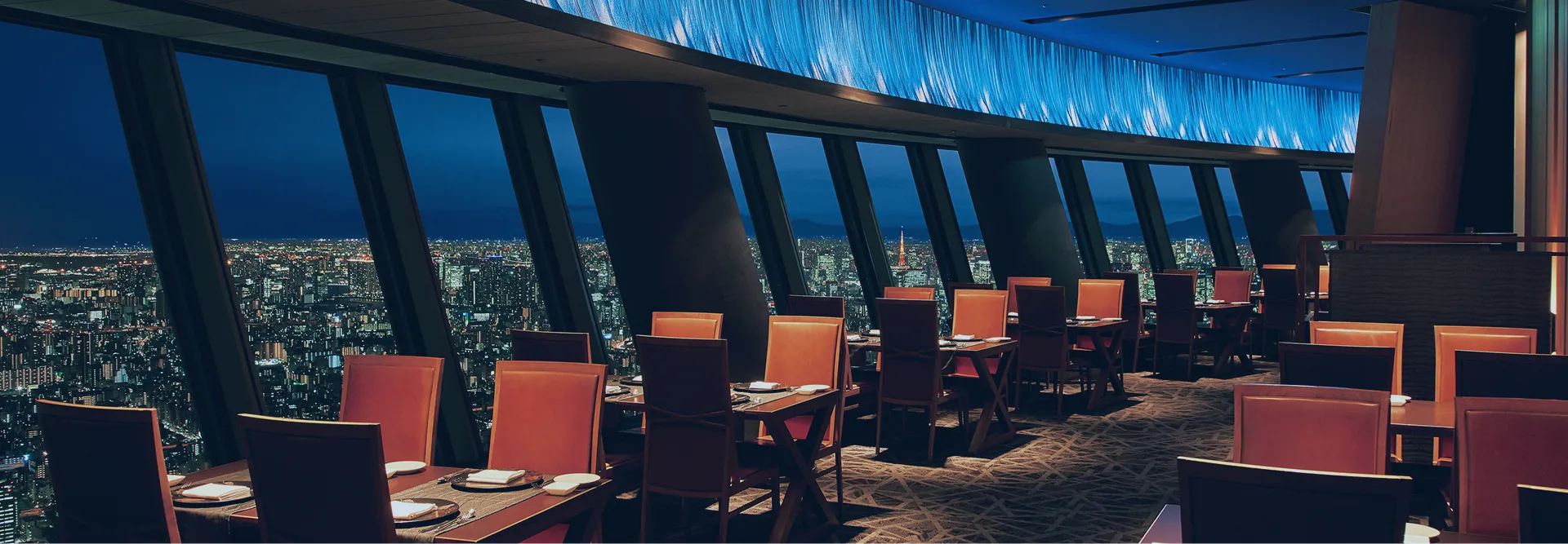 夜の街並みを一望できる、展望レストランの店内。大きな窓の向こうには東京の夜景が広がり、落ち着いた照明の中にテーブルセットが整然と並んでいます。
