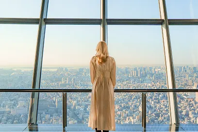 高層階の展望台で、大きな窓越しに広がる街の風景を背中で見つめる女性の後ろ姿