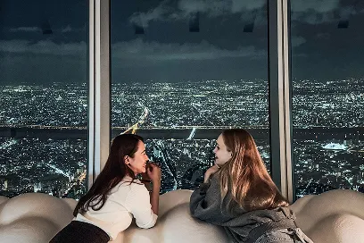 夜の展望台で、煌めく街の夜景が広がる窓際に並んで寄りかかり、微笑み合う2人の女性