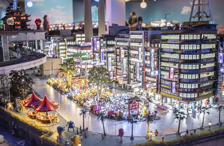 夜の街並みを再現した精巧なミニチュアジオラマ。ビルや屋台に明かりが灯り、小さな人々のフィギュアや乗り物が並ぶ活気ある風景。